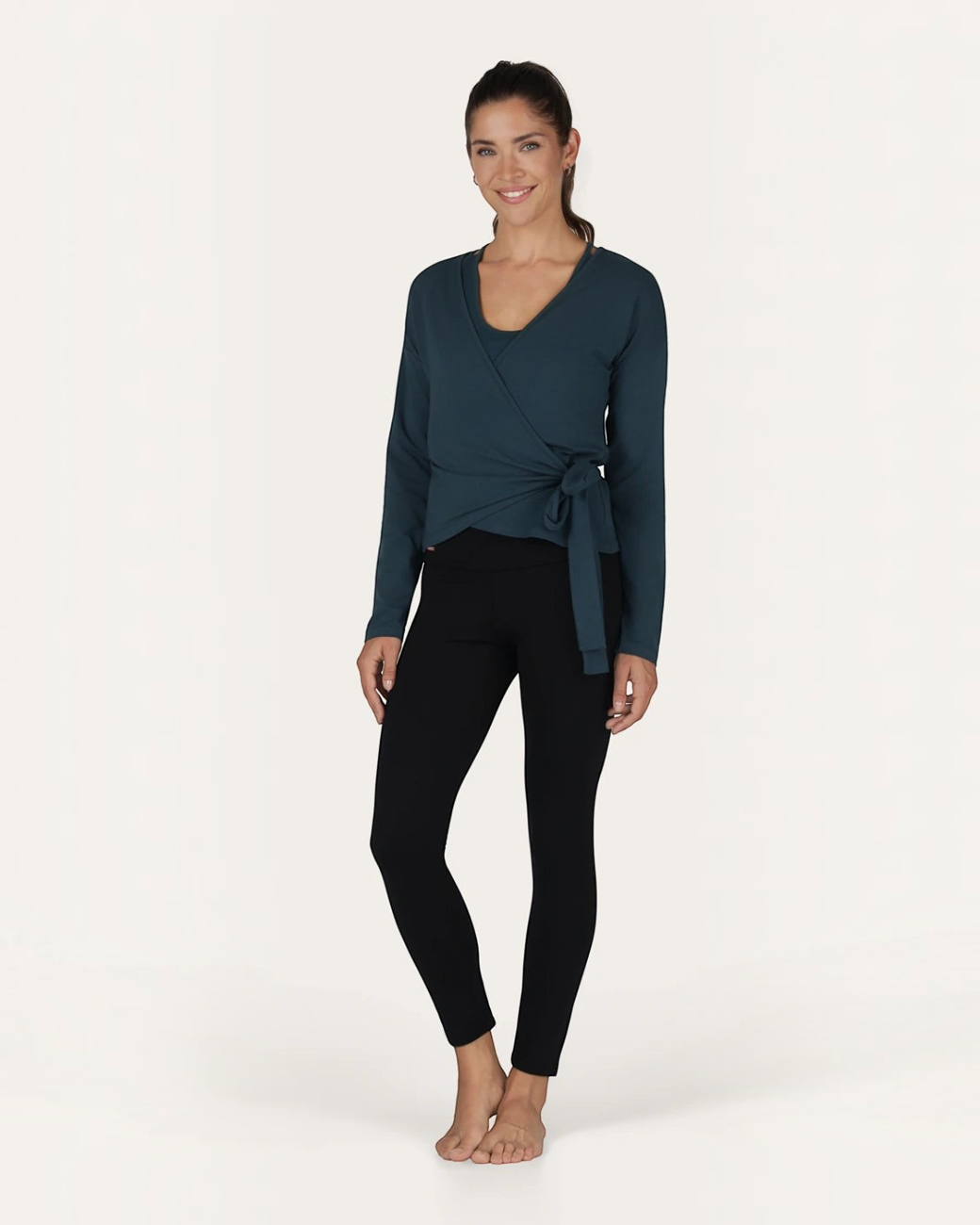 Model trägt grüne greenyogashop Aurora Wickeljacke aus Bambus-Mix mit Bindeband über einem Yoga-Top und Leggings